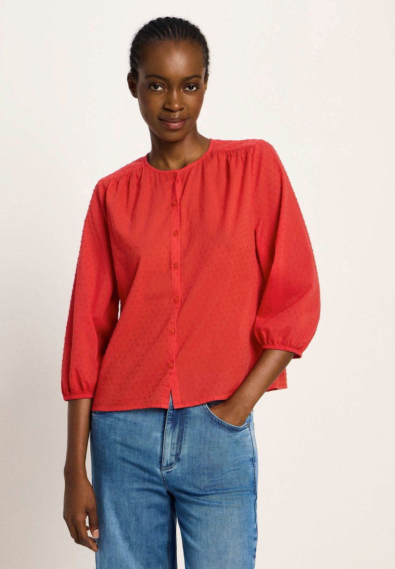 Rote Bluse mit Knopfverschluss und rundem Ausschnitt, strukturierter Stoff und dreiviertel Puffärmel, kombiniert mit blauen Jeans.