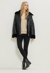 Giubbotto in pelle sintetica nera con colletto e polsini in pelliccia nera, abbinato a un maglione beige, jeans skinny neri e stivaletti neri.