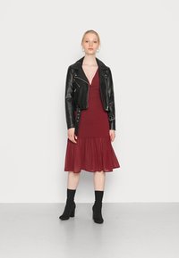 Casaco de cabedal preto tipo biker sobre um vestido burgundy profundo com bainha em bolsa, combinado com botas pretas até ao tornozelo e meias de malha.