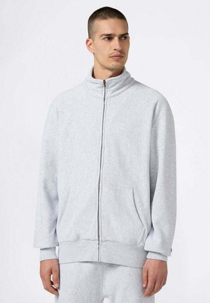 Mann in hellgrauem Sweatshirt mit Reißverschluss und passender Jogginghose, stehend vor weißem Hintergrund, blickt zur Seite.