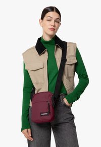 Borsa a tracolla Eastpak color bordeaux con chiusura a zip, dotata di una tasca frontale e tracolla regolabile, indossata sopra un gilet beige e un maglione verde.