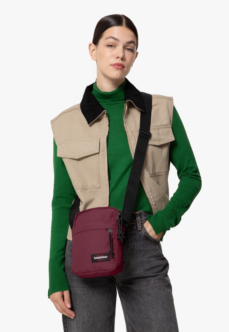Borsa a tracolla Eastpak color bordeaux con chiusura a zip, dotata di una tasca frontale e tracolla regolabile, indossata sopra un gilet beige e un maglione verde.