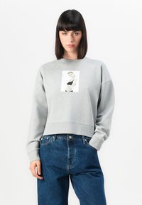 Donna con capelli scuri e lisci che indossa una felpa grigia con una toppa fotografica in bianco e nero, abbinata a jeans blu a vita alta, su sfondo bianco.