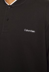 Camisa polo negra con un acabado texturizado, que presenta una tapeta con dos botones y el logotipo bordado en blanco "Calvin Klein" en el pecho.