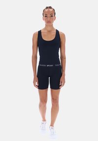 Sötétkék sportoverál testhezálló szabással, kerek nyakkivágással és hozzáillő rövidnadrággal. Fehér, márkázott elasztikus derékpánttal rendelkezik. Fehér tornacipővel viselve.