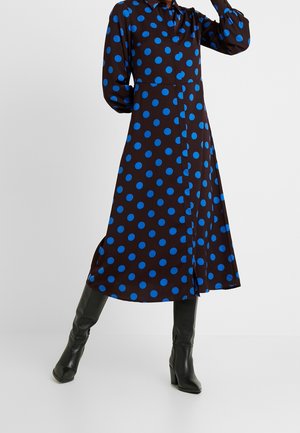 Woman wearing a black midi dress with blue polka dots and long puffed sleeves, paired with black heeled knee-high boots.