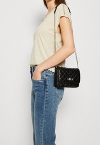 Quilted sort læder crossbody taske med en sølv kædetras, der har en frontlåg og metallukning, båret sammen med blå jeans og en lys top.