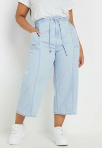 Pantalones culotte de denim azul claro con cintura ajustable, bolsillos laterales y detalles de costura visibles, combinados con zapatillas blancas.