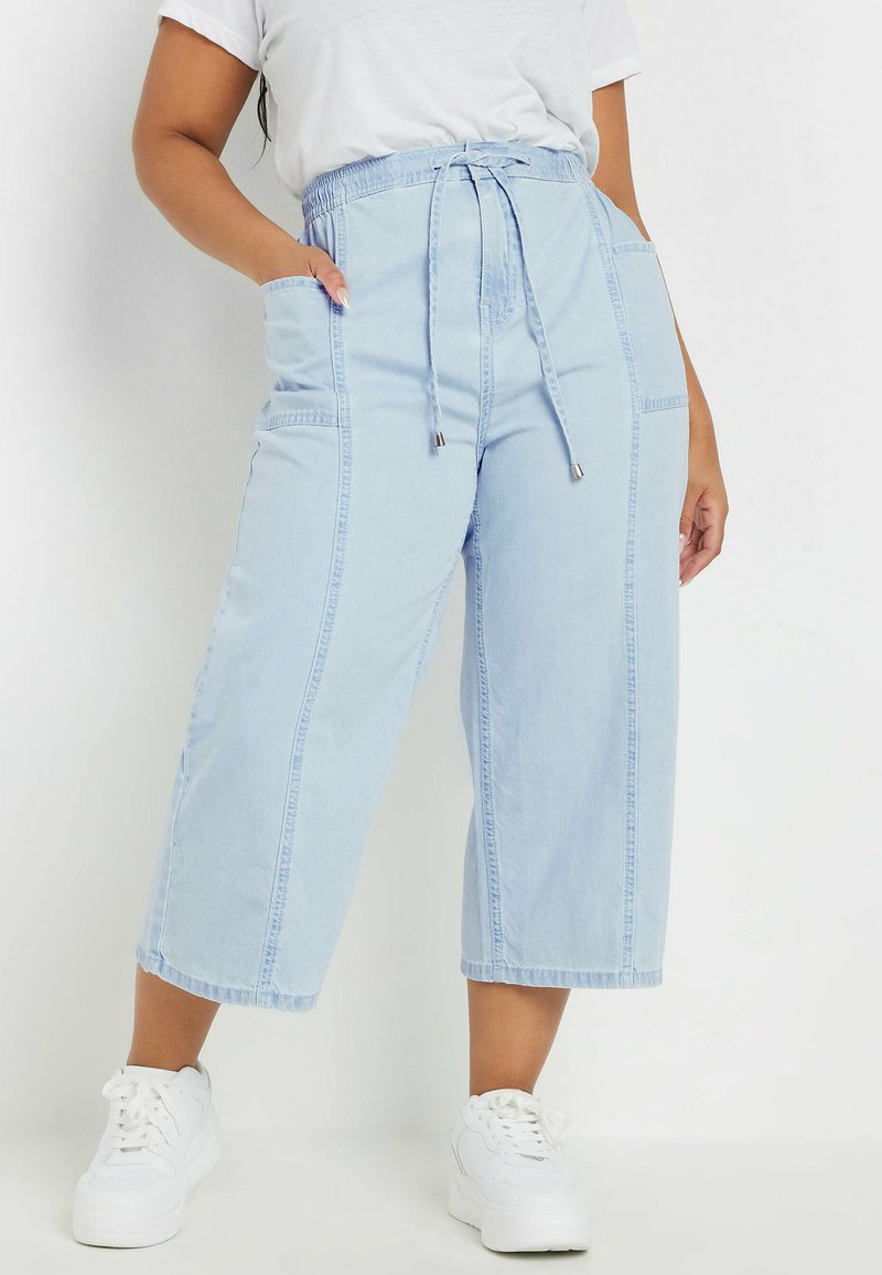 Pantalones culotte de denim azul claro con cintura ajustable, bolsillos laterales y detalles de costura visibles, combinados con zapatillas blancas.