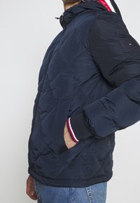 Man med mörkblå vadderad jacka och hand i fickan, randig manschettdetail och ljusblå jeans mot en enfärgad bakgrund.