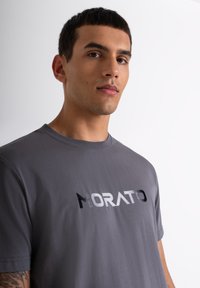 Siva pamučna majica s odvažnim logotipom "MORATO" u različitim nijansama, klasičnog okruglog izreza i kratkim rukavima. Glatka tekstura.