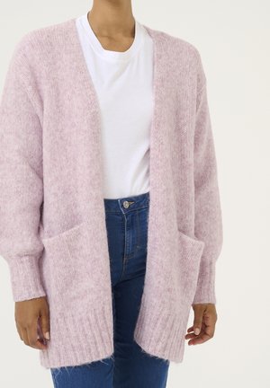 Kvinde iført lyserød, blød åben cardigan, hvid t-shirt og højtaljede blå jeans, med hænder afslappet langs siderne.
