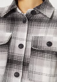 Black and white plaid shirt featuring a collar, black buttons, and two front pockets. Made of textured fabric with a checkered pattern.