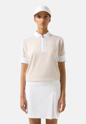 Vrouw met een beige korte mouwen polo, witte geplooide rok en witte pet, staand tegen een effen lichte achtergrond.