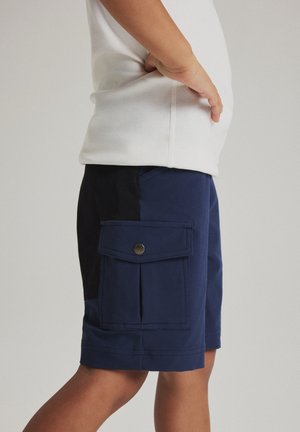 Kind draagt marineblauwe cargoshorts met klepzak en drukknoop, gecombineerd met een wit shirt, staand opzij.