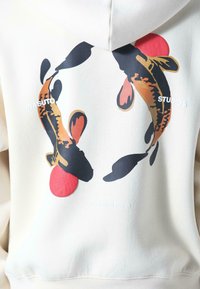 Crème hoodie met een achtergraphic van twee koi-vissen in oranje en zwart, vergezeld van rode cirkelaccenten en witte tekst.