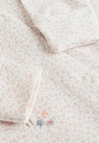 White fabric shirt with pink floral print, featuring button details, soft texture, and small embroidered accents in pink, blue, and beige.