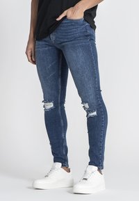 Jeans ajustados de mezclilla azul, con desgastes y rotos en las rodillas. Llevados con zapatillas blancas y una blusa negra.