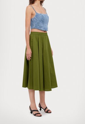 Femme portant une jupe midi verte, un crop top en denim bleu à fines bretelles, et des sandales noires à talon bas et bout ouvert, debout de profil.