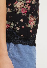 Top negro con estampado de rosas y hojas, con un borde de encaje en el dobladillo. Combinado con pantalones de pana azules.