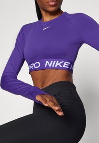 Lilla langermet crop topp med Nike-logo og bred elastikkant. Tettsittende passform, myk tekstur, kombinert med svarte høytliv leggings.