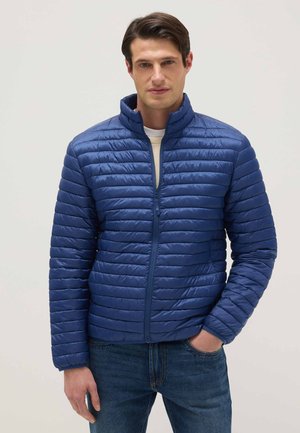 Hombre con chaqueta acolchada azul con cremallera y cuello alto, camiseta blanca debajo y vaqueros azules, de pie con una mano en el bolsillo.