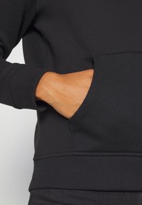 Mano con piel morena insertada en el bolsillo frontal de una sudadera negra con puños y bajo de canalé.
