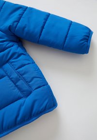 Szczegóły rękawa i kieszeni jasnoniebieskiej pikowanej kurtki na jasnoszarym tle.