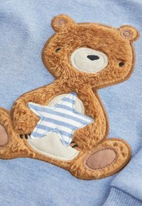Blauem Stoff mit einem gestickten Bären und einer flauschigen Textur, der einen gestreiften Stern hält. Der Bär hat braune und weiße Akzente.