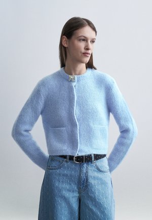 Donna che indossa un cardigan azzurro chiaro morbido con una fibbia a fiocco al collo e jeans azzurri a vita alta con una cintura nera, in piedi con le mani dietro la schiena.