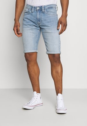 Man die lichtblauwe denim shorts met opgerolde zomen en witte hoge sneakers draagt, staand tegen een effen achtergrond.