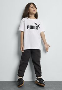 Valkoinen puuvillapaita, jossa on musta "PUMA"-logo, yhdistetty mustiin verkkareihin. Asusteena mustat lenkkarit, joissa on vaaleanruskeat pohjat.