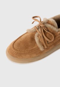 Bruine suède mocassin met beige bontvoering en veters, met een gestikt ontwerp en rubberen zool op een witte achtergrond.