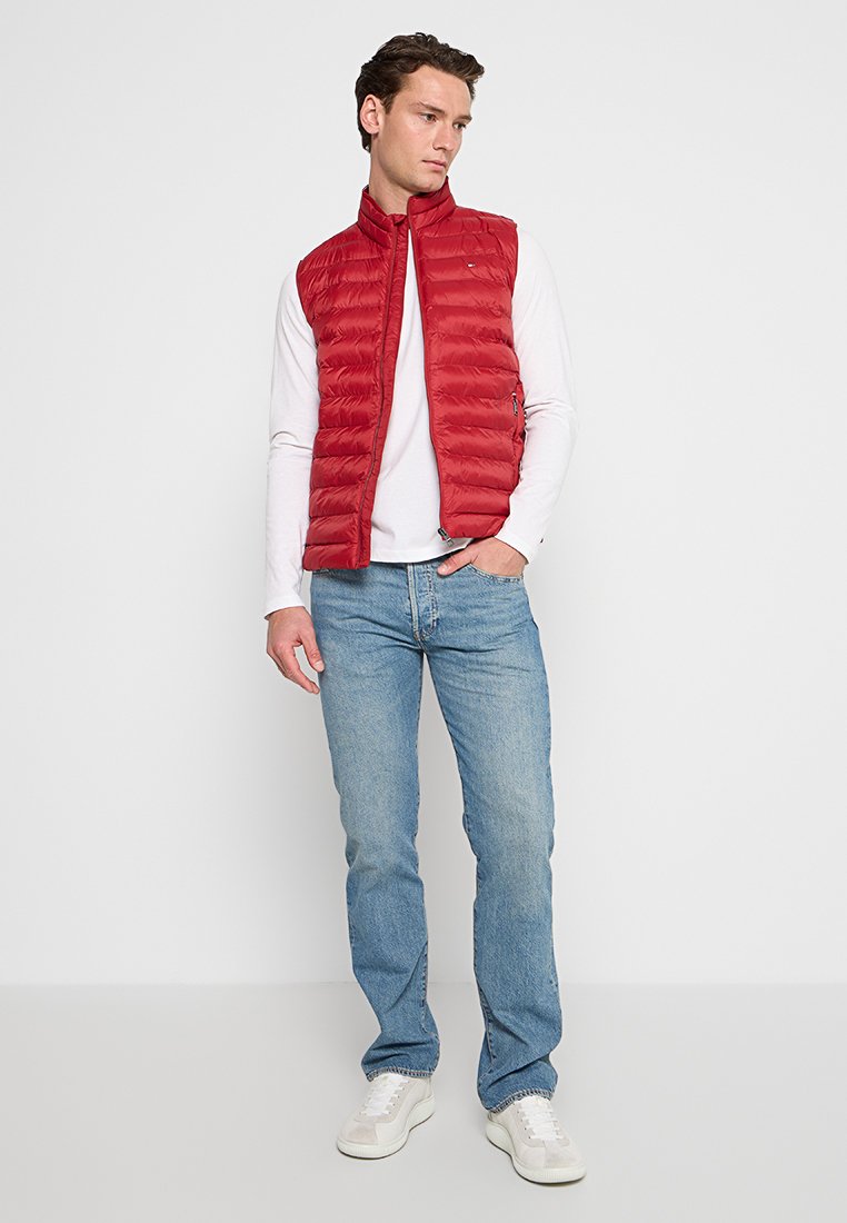 Gilet imbottito rosso con colletto alto, motivo a trapunta orizzontale; indossato sopra una camicia bianca a maniche lunghe, abbinato a jeans blu e sneakers bianche.