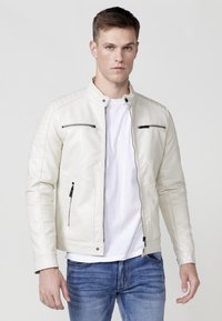 Chaqueta de cuero sintético blanco con cuello alto, que presenta dos bolsillos en el pecho con cremallera y acentos acolchados en los hombros. Llevada sobre una camisa blanca.