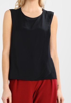 Sleeveless black blouse with round neckline worn with loose red pants on a standing person against a plain background.