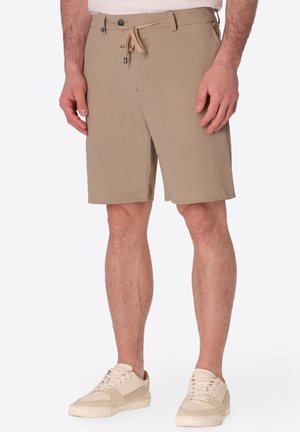 Uomo che indossa pantaloncini beige al ginocchio con coulisse in vita e sneakers avorio, in piedi davanti a uno sfondo semplice.