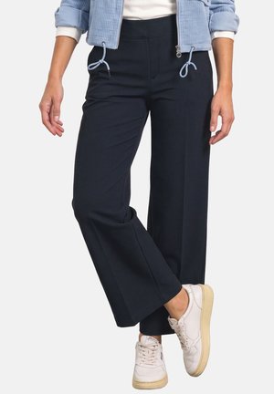 Personne portant un pantalon noir à jambes larges, des baskets blanches avec des semelles beige et une veste bleu clair avec les manches retroussées.