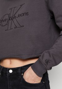 Izgriezts pelēks džemperis, izgatavots no mīksta auduma, ar ieplūdušu Calvin Klein Jeans logotipu. Piedurkņu maliņas ir ar ribotu apdari, un apakšā tas ir nedaudz izliekts.
