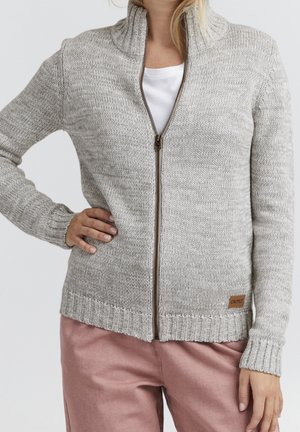 Kobieta ubrana w jasnoszary, dziergany sweter na zamek, na białym topie i różowych spodniach, ręka na biodrze, stoi na tle jednolitego tła.