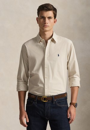 Beežist ja koorevärvi triibuline nööbitav särk rullitud varrukatega, millel on väike sinine logo. Kandatud tumedate teksade ja pruuni vööga.