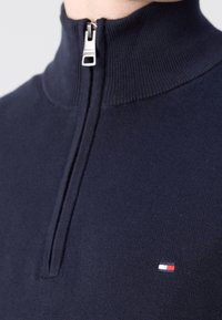 Pull à fermeture éclair bleu marine avec un col montant et un petit logo rectangulaire rouge, blanc et bleu sur la poitrine.
