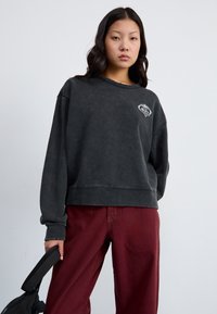 Felpa nera oversize con una texture leggermente vissuta e un logo a forma di cuore bianco sul petto, abbinata a pantaloni bordeaux scuri.
