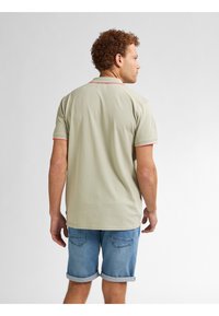 Lichtgroene polo met rode accenten op de kraag en mouwen, gecombineerd met opgerolde denimshorts. De stof lijkt glad.