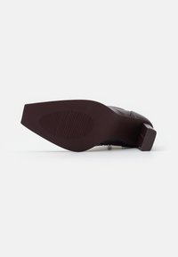 Zapato de tacón liso, de color marrón oscuro, tumbado de lado, mostrando suela suave con agarre ovalado texturizado y punta de tacón cuadrada.