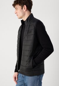 Colete acolchoado preto feito de tecido resistente à água; apresenta um fecho zip, gola alta e bolsos laterais para armazenamento. Design casual.