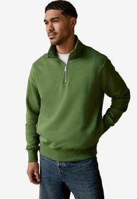 Green fleece pullover with a quarter-zip collar, ribbed cuffs, and a smooth texture, worn over a white t-shirt and denim jeans.