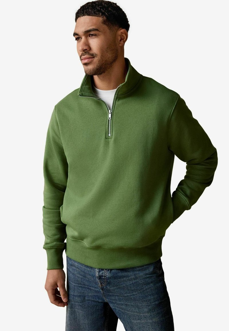 Green fleece pullover with a quarter-zip collar, ribbed cuffs, and a smooth texture, worn over a white t-shirt and denim jeans.