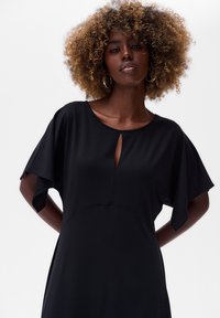 Schwarzes Kleid mit Rundhalsausschnitt, kurzen, flatternden Ärmelchen und einem Schlitzdetail vorne. Glattes Material und taillierte Passform.