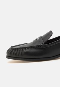 Mocassin en cuir noir avec des détails cousus et un design classique à bride penny sur une semelle plate, sur un fond blanc uni.
